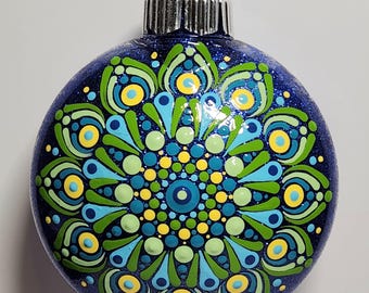 May include: A round, navy blue Christmas ornament with a glittery finish. The ornament features a hand-painted mandala design in shades of blue, green, and yellow. The design is symmetrical and detailed, with a silver metal cap and a black hanging loop.