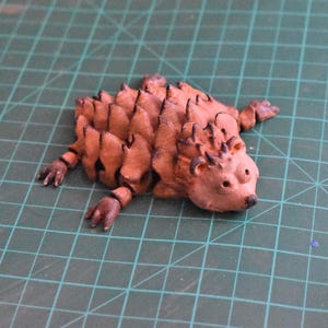 May include: A handcrafted, brown hedgehog figurine with detailed quills and small, articulated legs. The figurine is made of a clay-like material and is positioned on a green grid surface.