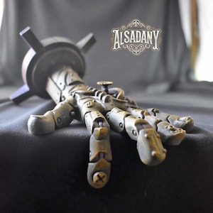 May include: A steampunk-style articulated hand made of metal and wood. The hand is painted in shades of brown, gold, and silver. The hand is attached to a base with a wooden platform and metal arms. The hand is posed with the fingers extended and the thumb pointing up. The hand is a unique and decorative piece of art.