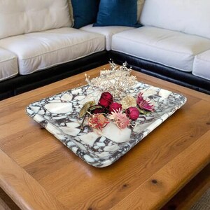 May include: A rectangular marble tray with a black and white pattern holds dried flowers and a decorative crown. The tray sits on a wooden coffee table with a lower shelf. Two blue velvet throw pillows rest on a white sectional sofa.