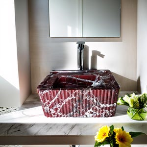 May include: A red marble bathroom sink with white veining, sitting on a white countertop. The sink is rectangular and has a rippled texture. A silver faucet is visible above the sink.