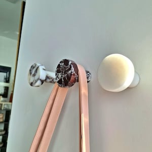 May include: Three decorative wall hooks. Two hooks feature a marble design with brown and white patterns, and one is solid white. A pink handbag strap hangs from one of the marble hooks.