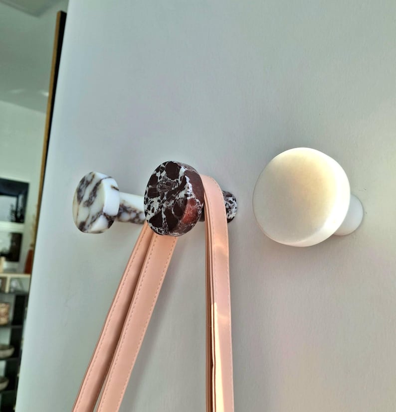 May include: Three decorative wall hooks. Two hooks feature a marble design with brown and white patterns, and one is solid white. A pink handbag strap hangs from one of the marble hooks.