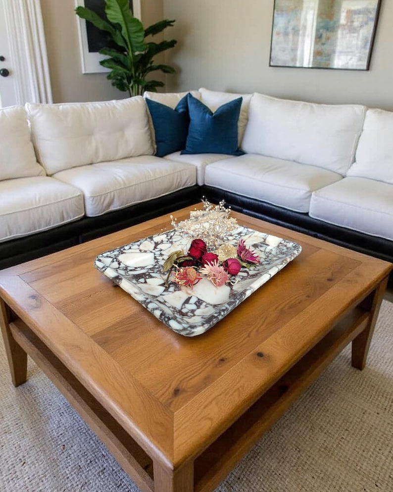 May include: A rectangular marble tray with a black and white pattern holds dried flowers and a decorative crown. The tray sits on a wooden coffee table with a lower shelf. Two blue velvet throw pillows rest on a white sectional sofa.