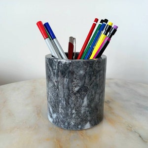May include: A grey marble pencil pot containing pens and pencils of various colours. The cylindrical holder sits on a marble surface. The pens and pencils include red, blue, and silver markers, and pencils with yellow, green, and pink accents.