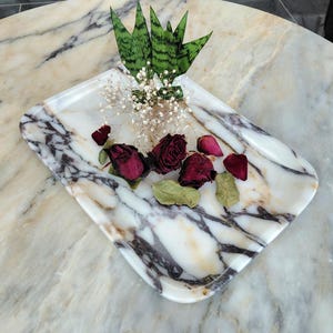 May include: A marble tray with a white and gray veined pattern, holding dried red roses, petals, and green leaves. The tray also features a small arrangement of green and white plants.