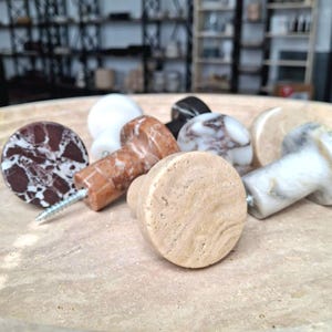May include: A selection of decorative cabinet knobs in various colours and patterns. The knobs are made of stone and marble, with a variety of textures and colours, including brown, white, and black. Each knob has a screw for easy installation.