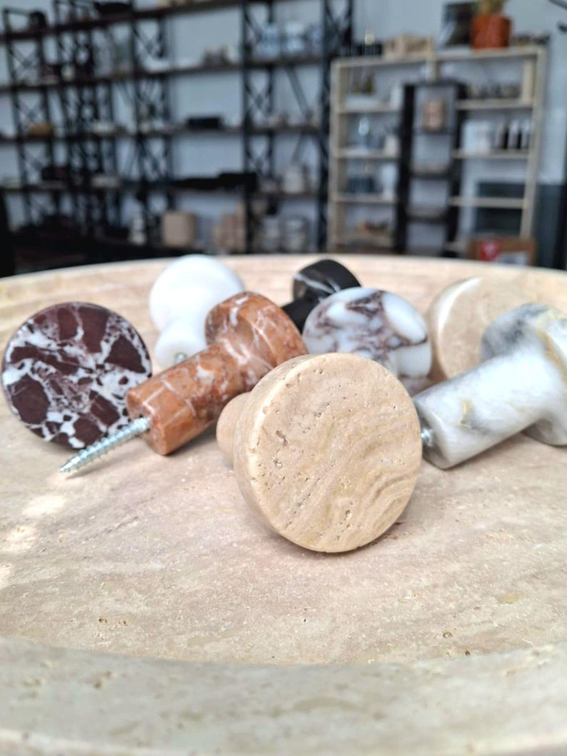 May include: A selection of decorative cabinet knobs in various colours and patterns. The knobs are made of stone and marble, with a variety of textures and colours, including brown, white, and black. Each knob has a screw for easy installation.