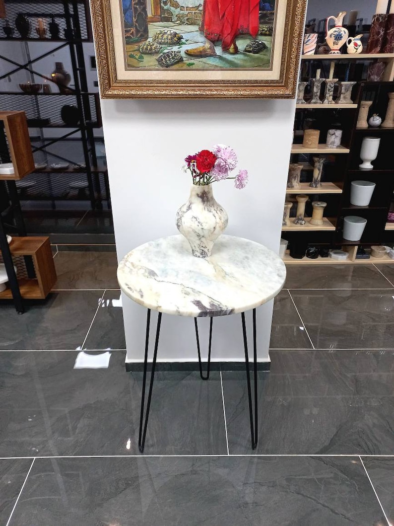 May include: A white marble top side table with black hairpin legs. A white ceramic vase with a pink and red flower arrangement sits on the table.