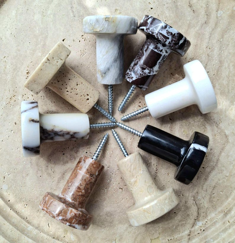 May include: A selection of eight decorative stone knobs in various colours and patterns, including white, black, brown, and beige. Each knob is attached to a silver screw, suitable for furniture or wall decor.