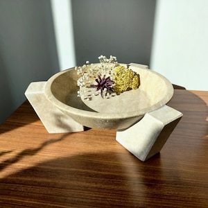 May include: A beige stone bowl with four legs sits on a wooden surface. The bowl is filled with dried flowers, including white and yellow blooms.