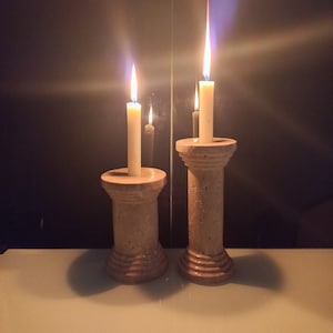 Könnte beinhalten: Zwei beigefarbene Kerzenhalter mit zylindrischem Design, jeder mit einer weißen Kerze mit einer brennenden Flamme. Die Kerzen stehen auf einer weißen Oberfläche.