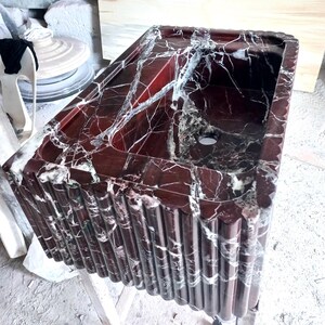 May include: A red and white marble bathroom sink with a unique fluted design. The sink is made of natural stone and has a smooth, polished finish.