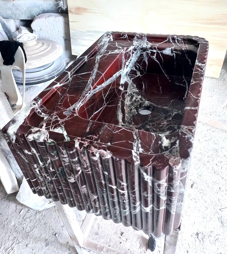 May include: A red and white marble bathroom sink with a unique fluted design. The sink is made of natural stone and has a smooth, polished finish.