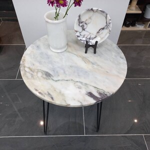 May include: A round white marble table with black hairpin legs. The table top has a light gray and white marbled pattern. A white ceramic vase with pink flowers and a white marble plate are on the table.