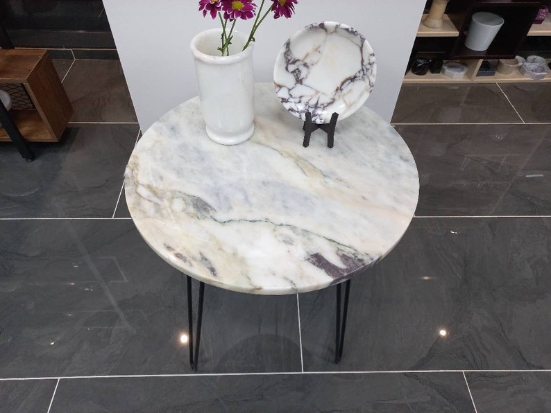 May include: A round white marble table with black hairpin legs. The table top has a light gray and white marbled pattern. A white ceramic vase with pink flowers and a white marble plate are on the table.