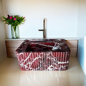 May include: A red marble sink with a white veining pattern. The sink has a rectangular shape with a rounded edge and a grooved design. A gold faucet is mounted above the sink.