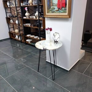 May include: A round marble table with a black metal base. The table is decorated with a white marble bowl and a vase of pink flowers. The table is in a room with a gray tiled floor and a shelf unit filled with pottery.