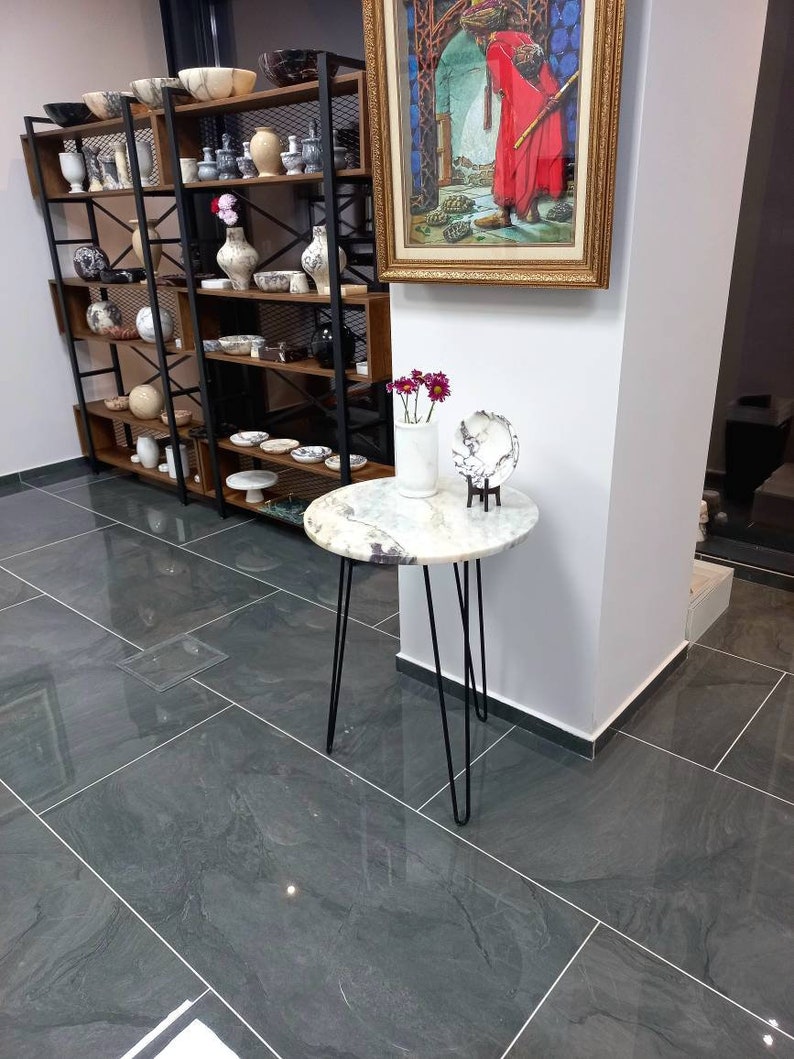 May include: A round marble table with a black metal base. The table is decorated with a white marble bowl and a vase of pink flowers. The table is in a room with a gray tiled floor and a shelf unit filled with pottery.