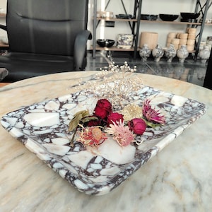 May include: A square marble tray with a white and purple pattern, holding dried flowers. The tray is on a marble surface. Shelves with various ceramic items are in the background.