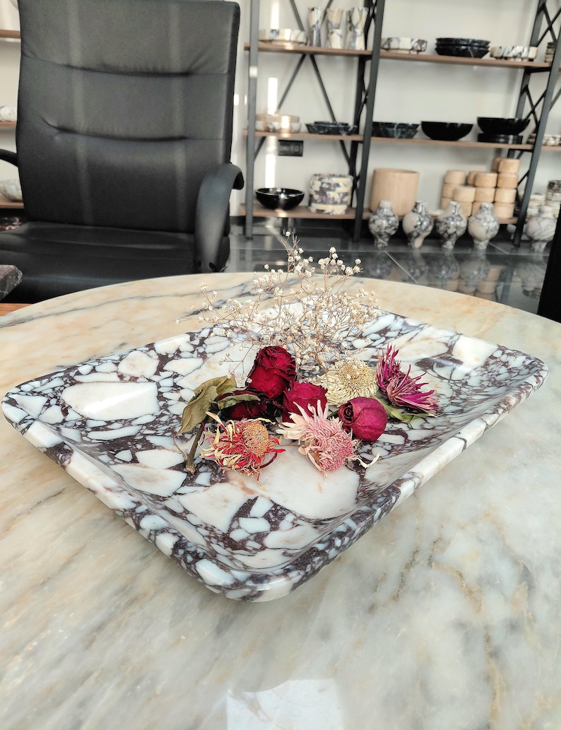 May include: A square marble tray with a white and purple pattern, holding dried flowers. The tray is on a marble surface. Shelves with various ceramic items are in the background.