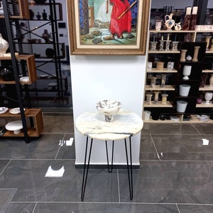 May include: A white marble round side table with black hairpin legs. The table is decorated with a white ceramic bowl with a decorative design.