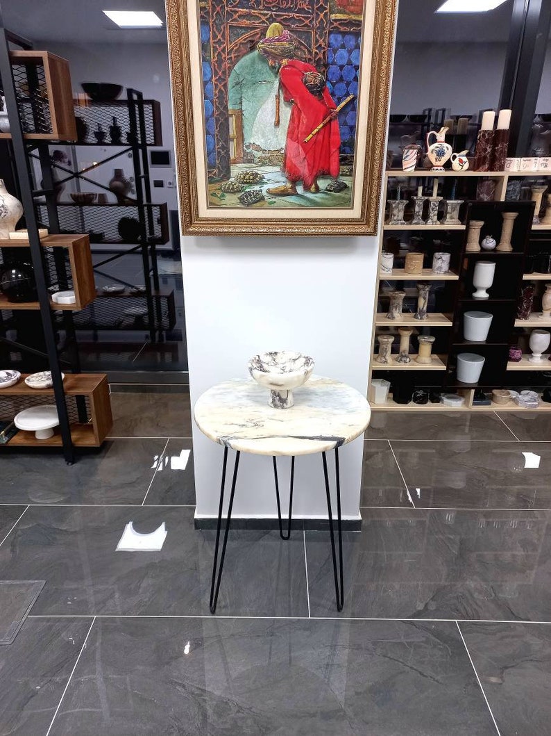 May include: A white marble round side table with black hairpin legs. The table is decorated with a white ceramic bowl with a decorative design.