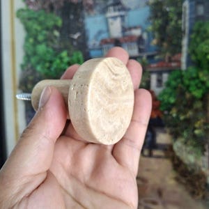 May include: A light beige, oval cabinet knob with a cylindrical handle. The knob is made of a stone-like material with a natural, textured appearance. A silver screw is attached to the handle for installation. The background is blurred.