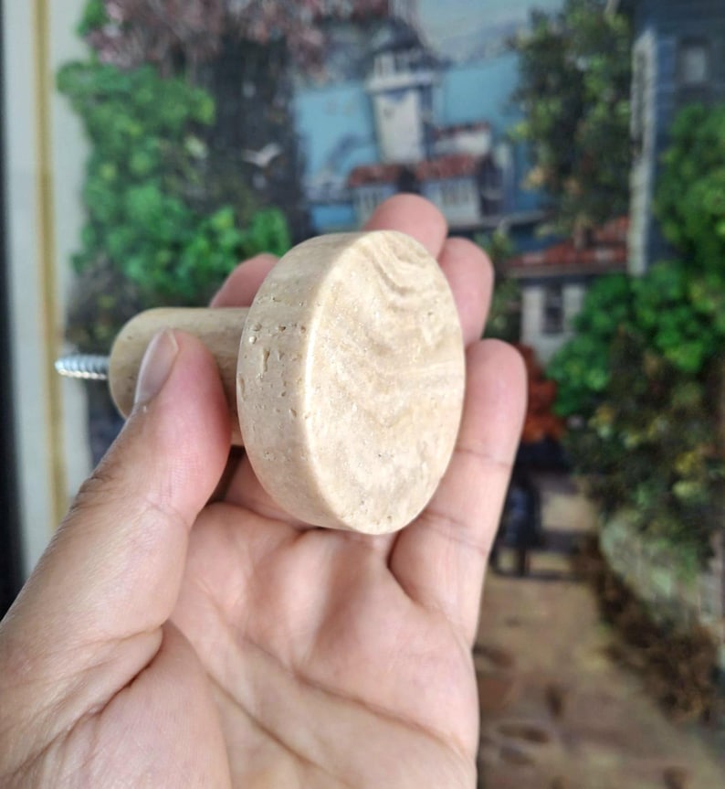 May include: A light beige, oval cabinet knob with a cylindrical handle. The knob is made of a stone-like material with a natural, textured appearance. A silver screw is attached to the handle for installation. The background is blurred.