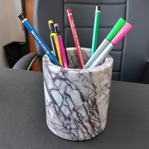 Puede incluir: Un porta lápices de mármol con un patrón de mármol blanco y gris. El porta lápices está lleno de lápices y bolígrafos de varios colores, incluyendo un lápiz azul, un lápiz rojo, un bolígrafo verde, un bolígrafo rosa y un lápiz amarillo.