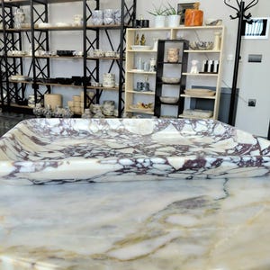 May include: A rectangular marble tray with a cream-colored base and intricate purple veining. The tray is displayed on a marble surface, showcasing its polished finish. Shelves filled with various ceramic items are visible in the background.