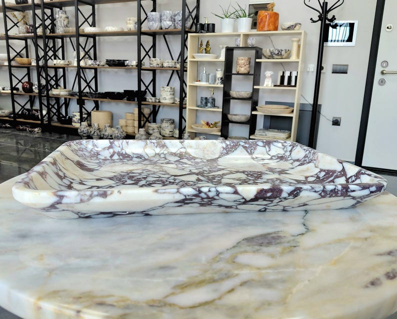 May include: A rectangular marble tray with a cream-colored base and intricate purple veining. The tray is displayed on a marble surface, showcasing its polished finish. Shelves filled with various ceramic items are visible in the background.