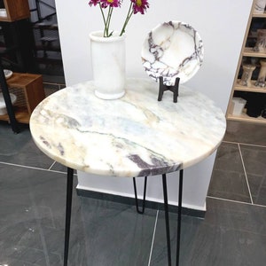 May include: A round marble table with black hairpin legs. The table top has a white, gray, and purple marbled pattern. A white vase with purple flowers sits on the table, and a white marble plate with a purple marbled pattern sits on a black stand.