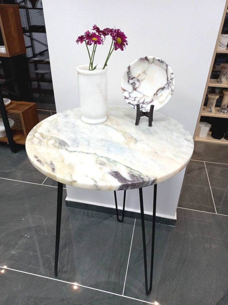 May include: A round marble table with black hairpin legs. The table top has a white, gray, and purple marbled pattern. A white vase with purple flowers sits on the table, and a white marble plate with a purple marbled pattern sits on a black stand.