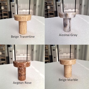 May include: Four stone bottle stoppers in beige travertine, Aizonai grey, Aegean rose, and beige marble. Each stopper has a cylindrical body and a round top. The names of the stones are printed below each stopper.