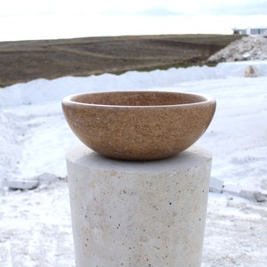 May include: A brown stone bowl sits on a white stone pedestal. The bowl is round and has a rough texture. The pedestal is tall and cylindrical.