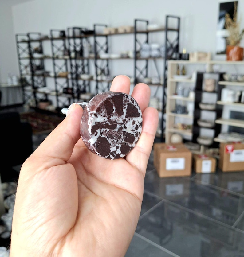May include: A circular stone object, predominantly dark brown with white veining. The object is held in a hand, highlighting its size and texture. The stone has a marbled appearance, with a mix of dark and light colours.