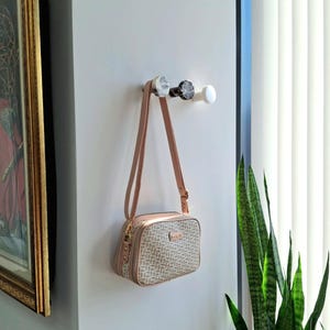 May include: A beige and brown crossbody bag with a repeating pattern hangs from a wall hook. The bag has a long, adjustable strap and gold-coloured hardware. Three decorative wall hooks are visible, with marble and white finishes.