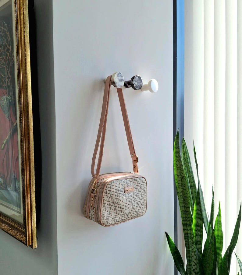 May include: A beige and brown crossbody bag with a repeating pattern hangs from a wall hook. The bag has a long, adjustable strap and gold-coloured hardware. Three decorative wall hooks are visible, with marble and white finishes.