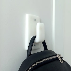 May include: White ceramic wall hook with a rounded hook shape. A black leather backpack is hanging on the hook.