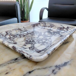 May include: Rectangular marble tray with a white and dark purple veined pattern. The tray has rounded corners and a slightly raised edge. The tray is sitting on a marble table. Home decor item.