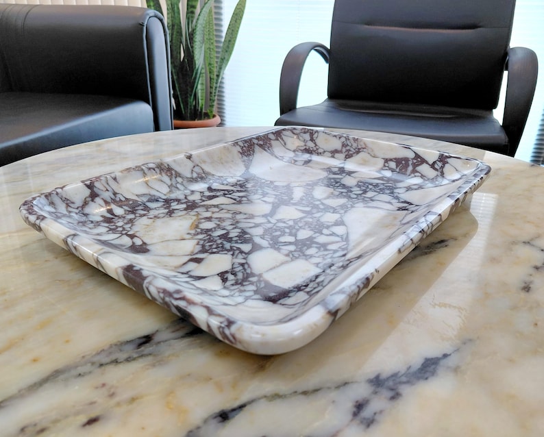 May include: Rectangular marble tray with a white and dark purple veined pattern. The tray has rounded corners and a slightly raised edge. The tray is sitting on a marble table. Home decor item.