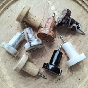 May include: A selection of stone knobs in diverse colours and patterns, such as beige, brown, black, white, and marble. Each knob features a cylindrical base and a screw for simple installation. These knobs are suitable for cabinets and drawers.