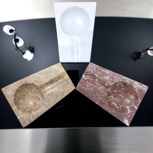 Puede incluir: Tres ceniceros de mármol, uno blanco, uno marrón y uno rosa, dispuestos en forma de triángulo sobre una superficie negra.