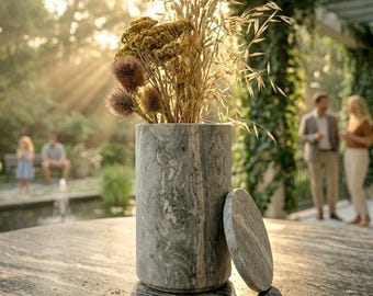 May include: A cylindrical gray marble container with a matching circular lid resting beside it. The container is filled with a bouquet of dried grasses and golden-brown wildflowers, featuring a distinct swirling pattern in the natural stone.