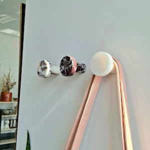 May include: Three decorative wall hooks. Two hooks feature a marbled pattern in white, black, and brown. The third hook is white and holds a pink strap. The hooks are mounted on a white wall.