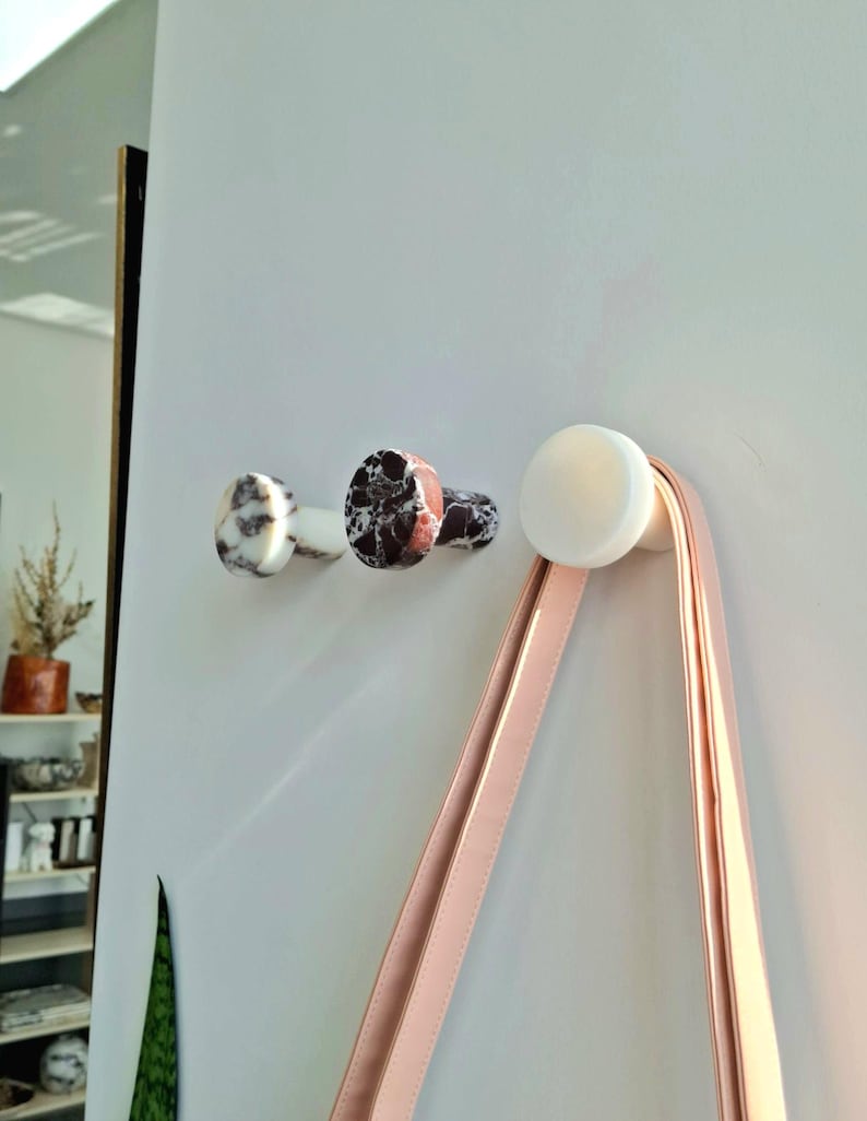 May include: Three decorative wall hooks. Two hooks feature a marbled pattern in white, black, and brown. The third hook is white and holds a pink strap. The hooks are mounted on a white wall.