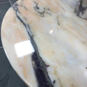 May include: A close-up of a round, white marble table top with a swirling, gray and black pattern. The surface is smooth and polished.
