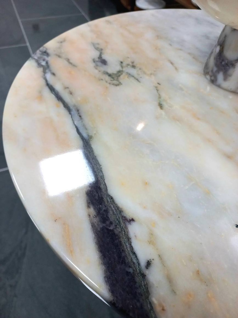 May include: A close-up of a round, white marble table top with a swirling, gray and black pattern. The surface is smooth and polished.