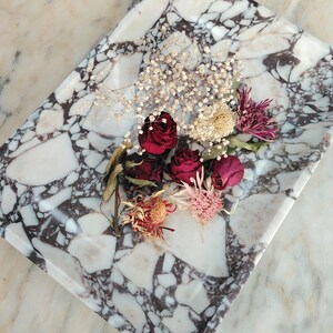 May include: A rectangular marble tray with a white and dark gray pattern, holding dried flowers. The flowers include deep red roses, small white blooms, and various dried flower heads in shades of pink and orange.